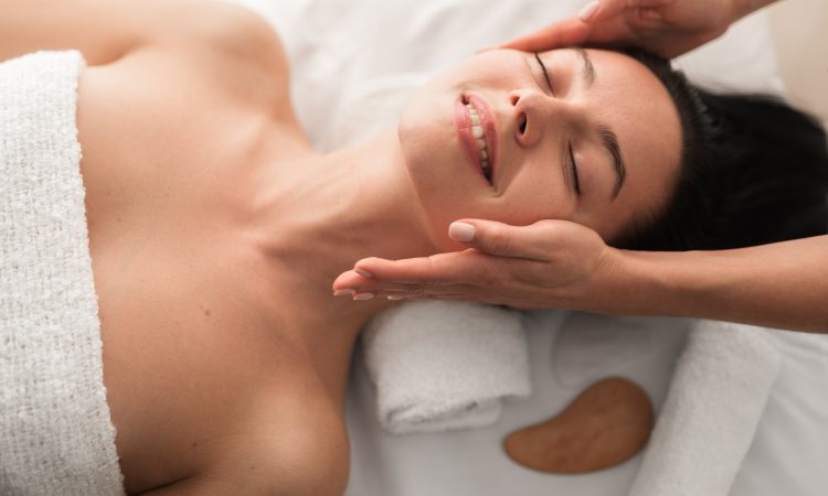 Top view of crop unrecognizable cosmetician doing face massage to cheerful young female customer with long dark hair, lying on couch with closed eyes and smiling